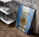 Argentina football national team crest jersey poster in a black frame leaning beside a shelf in a raw-plaster loft