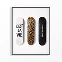 Three Saint Laurent Cest La Vie skateboards displayed in a black-framed poster: white with handwritten text, leopard print.