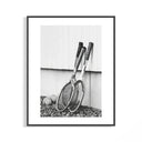 Black and white photo of vintage wooden tennis rackets leaning on a white wall over rocky ground