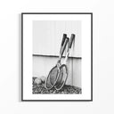 Black and white photograph of vintage wooden tennis rackets leaning against a white wall on rocky ground.