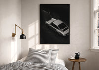Silver Porsche 911 parked print in a black frame above a bed with striped linens, a brass wall lamp and a sunlit window