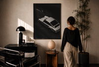 Silver Porsche 911 parked in alley print in a black frame on a sunlit plaster wall, a woman looking on beside a mushroom lamp