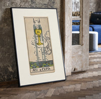 Ace of Cups tarot card print in a black frame leaning beside a vintage door on parquet