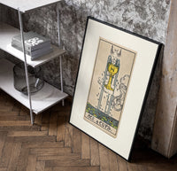 Ace of Cups tarot card poster in a black frame leaning beside a shelf in a raw-plaster loft