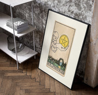 Hand holding pentacle coin poster in a black frame leaning beside a shelf in a raw-plaster loft