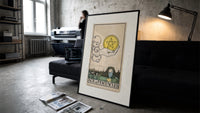 Hand holding pentacle coin print in a slim black frame leaning on a dark sofa in a concrete print studio