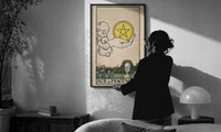 Ace of Pentacles print in a slim black frame being hung on a sunlit wall in a monochrome room