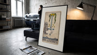 Hand holding upright sword with crown print in a slim black frame leaning on a dark sofa in a concrete print studio