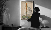 Ace of Wands print in a slim black frame being hung on a sunlit wall in a monochrome room