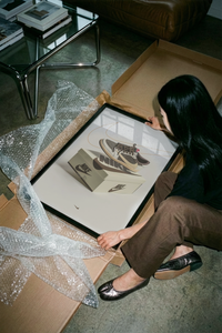 Woman unboxing framed Air Jordan 1 Travis Scott Reverse Mocha artwork from cardboard packaging on concrete floor