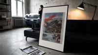 Alpine mountain and lake landscape print in a slim black frame leaning on a dark sofa in a concrete print studio
