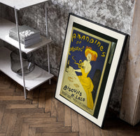 Woman in yellow eating biscuit poster in a black frame leaning beside a shelf in a raw-plaster loft