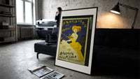 Woman in yellow eating biscuit print in a slim black frame leaning on a dark sofa in a concrete print studio