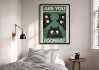 Are You Pooping Three Cats print in a black frame above a bed with striped linens, a brass wall lamp and a sunlit window