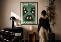 Are You Pooping Three Cats print in a black frame on a sunlit plaster wall, a woman looking on beside a mushroom lamp