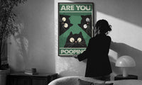 Are You Pooping Three Cats print in a slim black frame being hung on a sunlit wall in a monochrome room