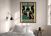 Are You Pooping Three Cows print in a black frame above a bed with striped linens, a brass wall lamp and a sunlit window