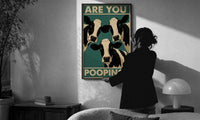 Are You Pooping Three Cows print in a slim black frame being hung on a sunlit wall in a monochrome room