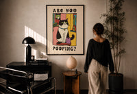 Are You Pooping Tuxedo Cat print in a black frame on a sunlit plaster wall, a woman looking on beside a mushroom lamp