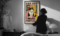 Are You Pooping Tuxedo Cat print in a slim black frame being hung on a sunlit wall in a monochrome room