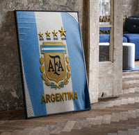 Argentina football national team crest jersey print in a black frame leaning beside a vintage door on parquet