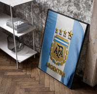 Argentina football national team crest jersey poster in a black frame leaning beside a shelf in a raw-plaster loft