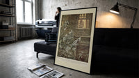 Ghostly phantoms floating outside window print in a slim black frame leaning on a dark sofa in a concrete print studio
