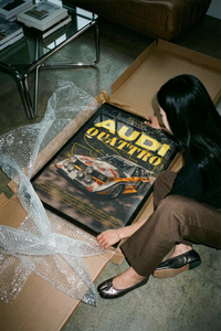 Woman lifting a framed Audi rally poster from a cardboard box with bubble wrap near a leather sofa