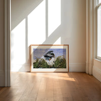 Great blue heron in marsh print in an oak frame on a sunlit wood floor beneath tall window light