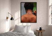 Back View Fastening a Necklace print in a black frame above a bed with striped linens, a brass wall lamp and a sunlit window