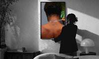 Back View Fastening a Necklace print in a slim black frame being hung on a sunlit wall in a monochrome room