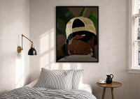 Backwards Cap with an Embracing Hand print in a black frame above a bed with striped linens