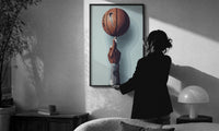 Basketball Balanced on a Fingertip print in a slim black frame being hung on a sunlit wall in a monochrome room