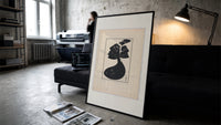 Woman in black gown and hat print in a slim black frame leaning on a dark sofa in a concrete print studio