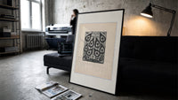 Peacock feather title page design print in a slim black frame leaning on a dark sofa in a concrete print studio