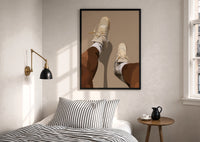 Beige Sneakers and White Socks print in a black frame above a bed with striped linens, a brass wall lamp and a sunlit window