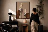Beige Sneakers and White Socks print in a black frame on a sunlit plaster wall, a woman looking on beside a mushroom lamp