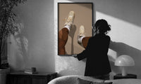 Beige Sneakers and White Socks print in a slim black frame being hung on a sunlit wall in a monochrome room