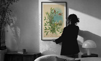 Birds Wishing You a Merry Christmas print in a slim black frame being hung on a sunlit wall in a monochrome room