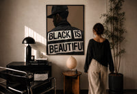 Black Is Beautiful Cap print in a black frame on a sunlit plaster wall, a woman looking on beside a mushroom lamp