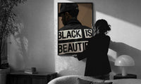 Black Is Beautiful Cap print in a slim black frame being hung on a sunlit wall in a monochrome room