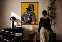 Black Is Beautiful Flame Jacket print in a black frame on a sunlit plaster wall, a woman looking on beside a mushroom lamp
