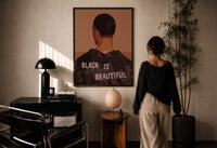 Black Is Beautiful Jacket print in a black frame on a sunlit plaster wall, a woman looking on beside a mushroom lamp