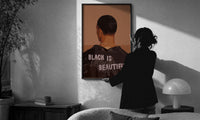 Black Is Beautiful Jacket print in a slim black frame being hung on a sunlit wall in a monochrome room