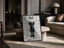 Black KAWS Companion figure standing print in a black frame leaning on a travertine table in a sunlit lounge