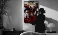 Black Man Having His Hair Braided print in a slim black frame being hung on a sunlit wall in a monochrome room