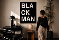 Black Man Lettering print in a black frame on a sunlit plaster wall, a woman looking on beside a mushroom lamp