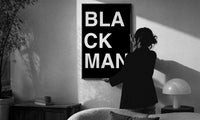 Black Man Lettering print in a slim black frame being hung on a sunlit wall in a monochrome room