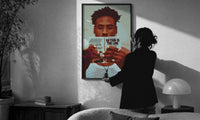 Black Man Reading a Paperback print in a slim black frame being hung on a sunlit wall in a monochrome room