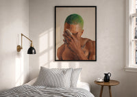 Black Man with Green Hair print in a black frame above a bed with striped linens, a brass wall lamp and a sunlit window